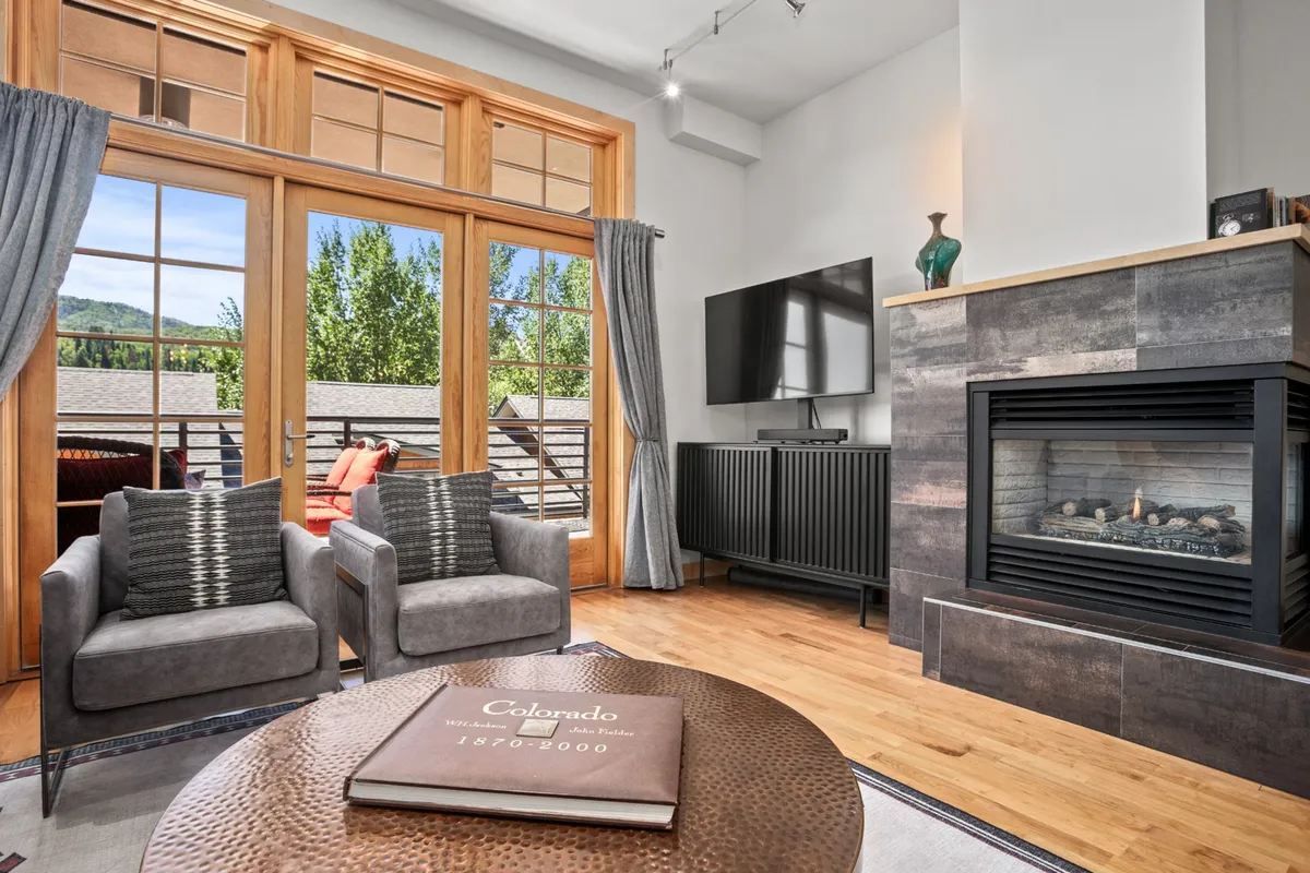 A welcoming living area featuring a comfortable seating arrangement around a fireplace, large windows with scenic views of the mountains, and a coffee table with a book. The setting is warm and perfect for relaxing or socializing, tying in both the mountain backdrop and the cozy interior elements.
