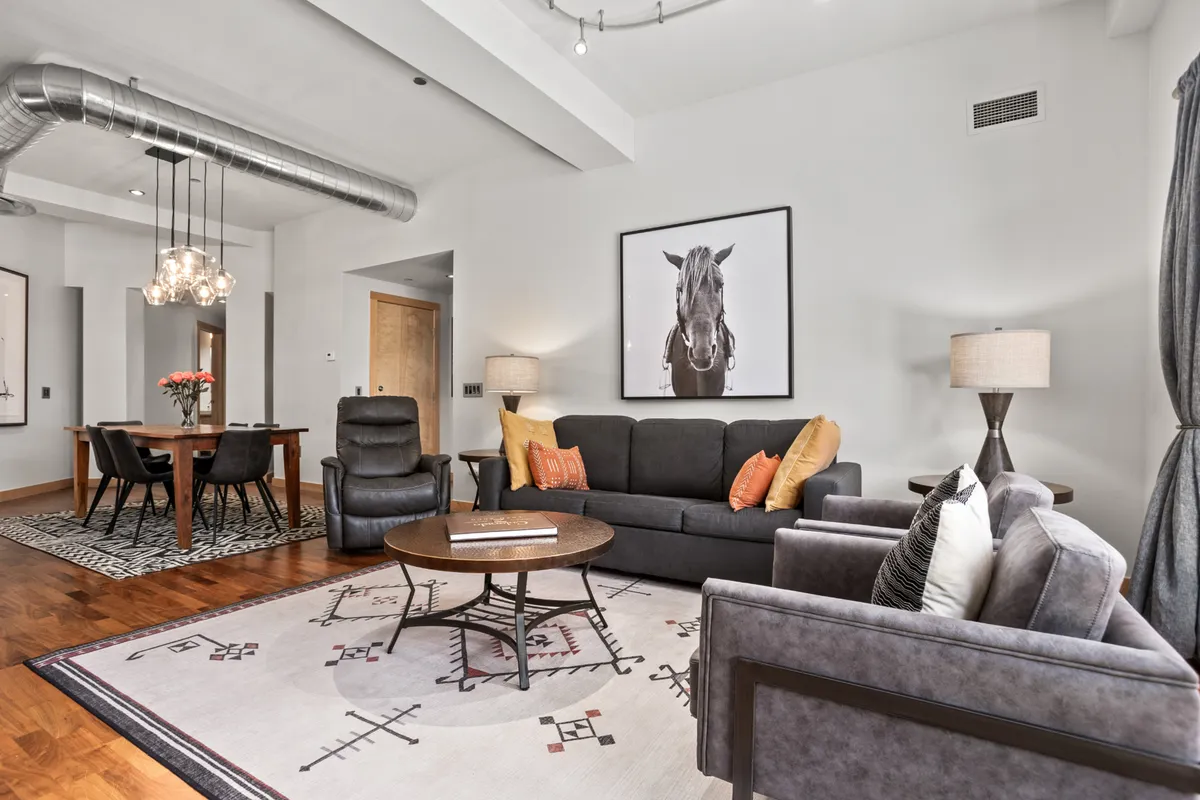 A comfortable living area featuring a stylish sofa with orange cushions, a contemporary dining table, striking horse artwork, and a modern floor lamp. The space exudes an inviting, modern vibe ideal for relaxation and small gatherings.