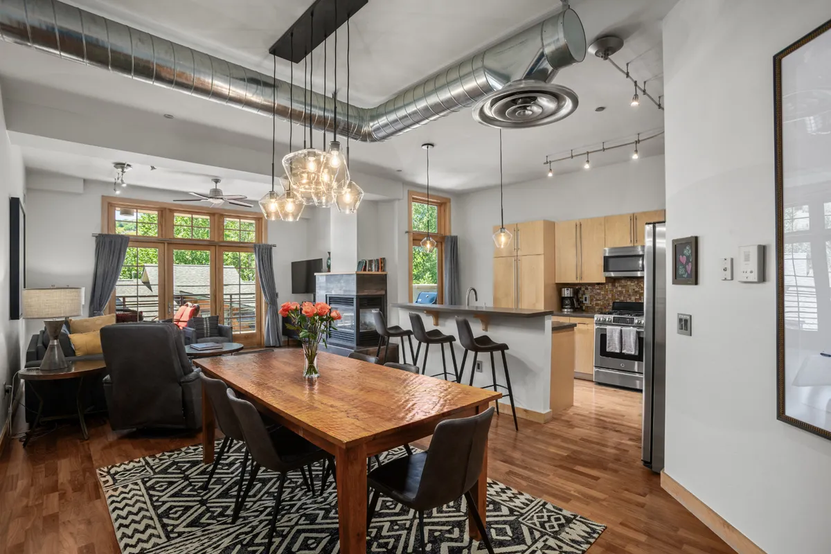 This image showcases a stylish dining table adorned with a vase of flowers, modern light fixtures overhead, an open kitchen layout, and large windows that allow natural light to flood the space. The arrangement highlights seamless flow between dining and kitchen areas, capturing the atmosphere of open-concept living and entertaining.