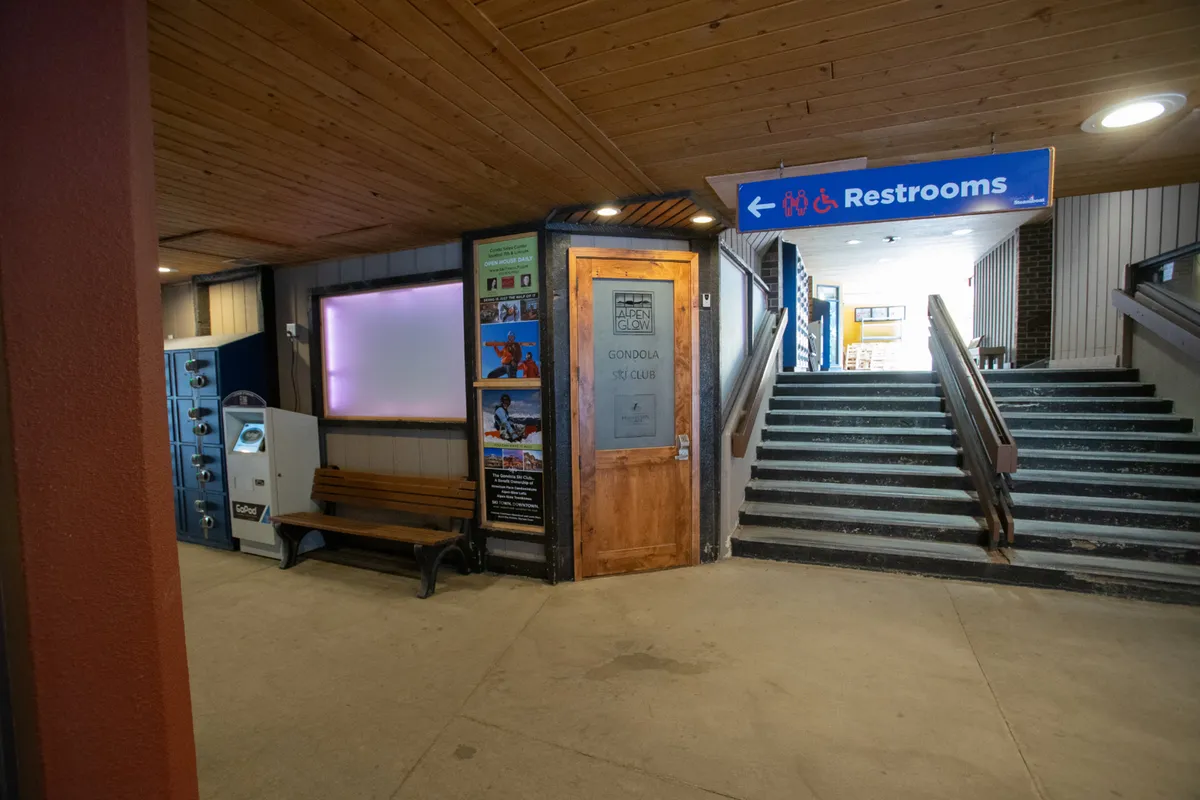 This image captures the entrance to the Gondola Ski Club, featuring the ski club door prominently, as well as benches, lockers, an information board, and visible ski-related signage. The functional yet welcoming space is set up for skiers to store their gear, take breaks, and plan their day on the slopes.