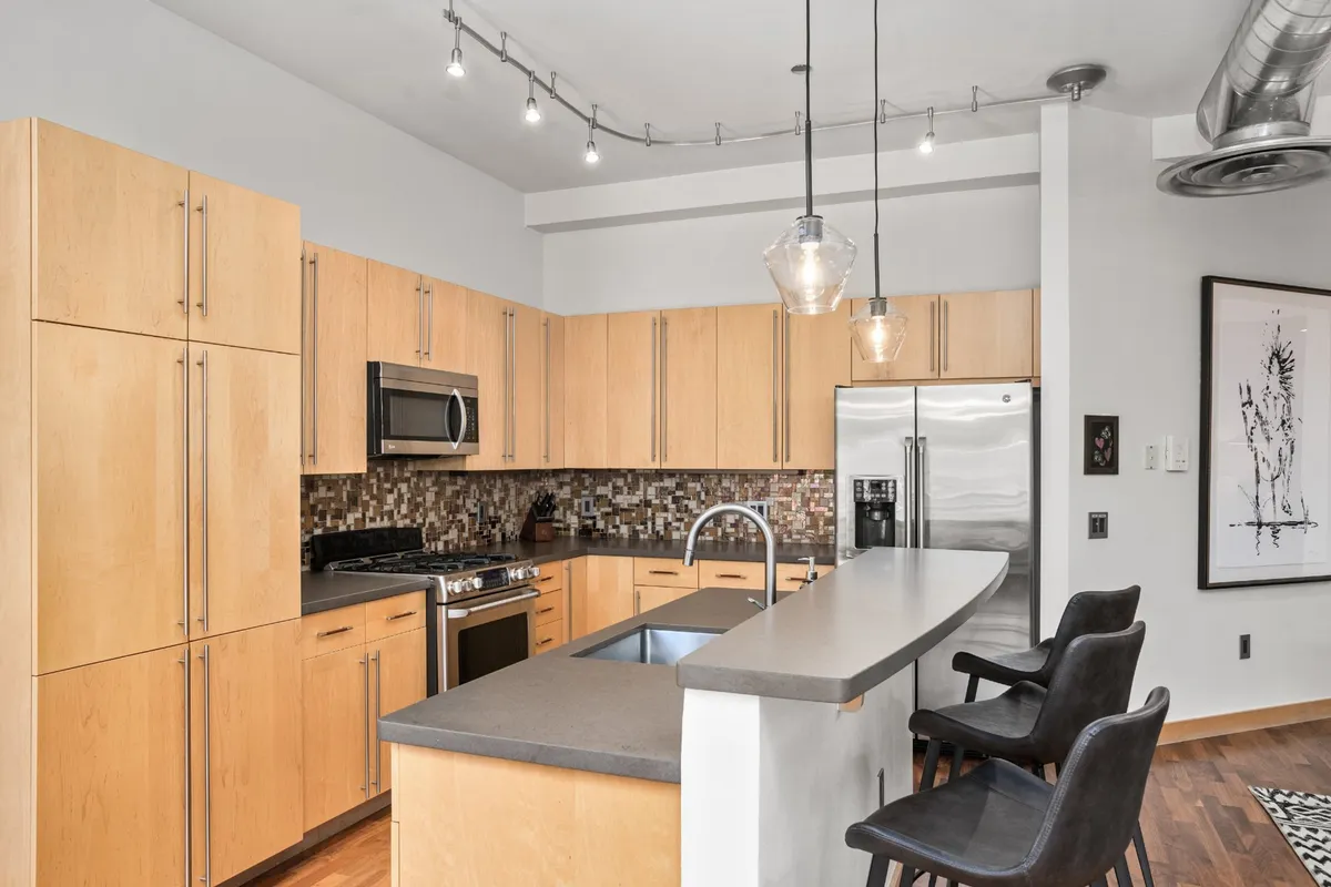 A modern kitchen with an island, stainless steel appliances, hanging light fixtures, and a decorative mosaic backsplash. The layout is open and inviting, designed for cooking and entertaining.