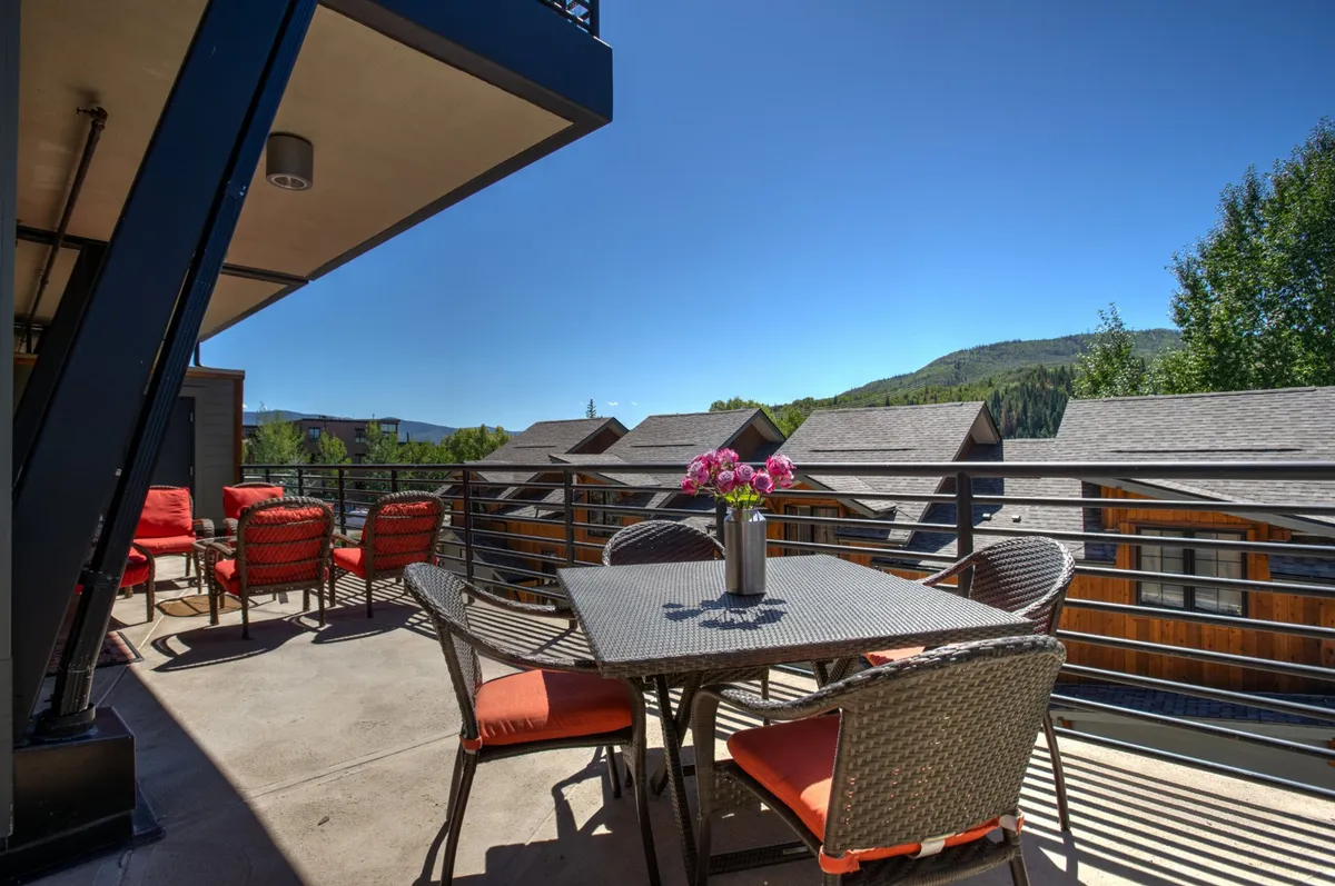 An inviting exterior scene showcasing a spacious outdoor dining table and comfortable seating area, set against a backdrop of mountains and vibrant, colorful flowers. The area is bathed in sunlight with ample space for relaxing, socializing, or enjoying a meal outside. The mountain view hints at the natural beauty and outdoor attractions close by.
