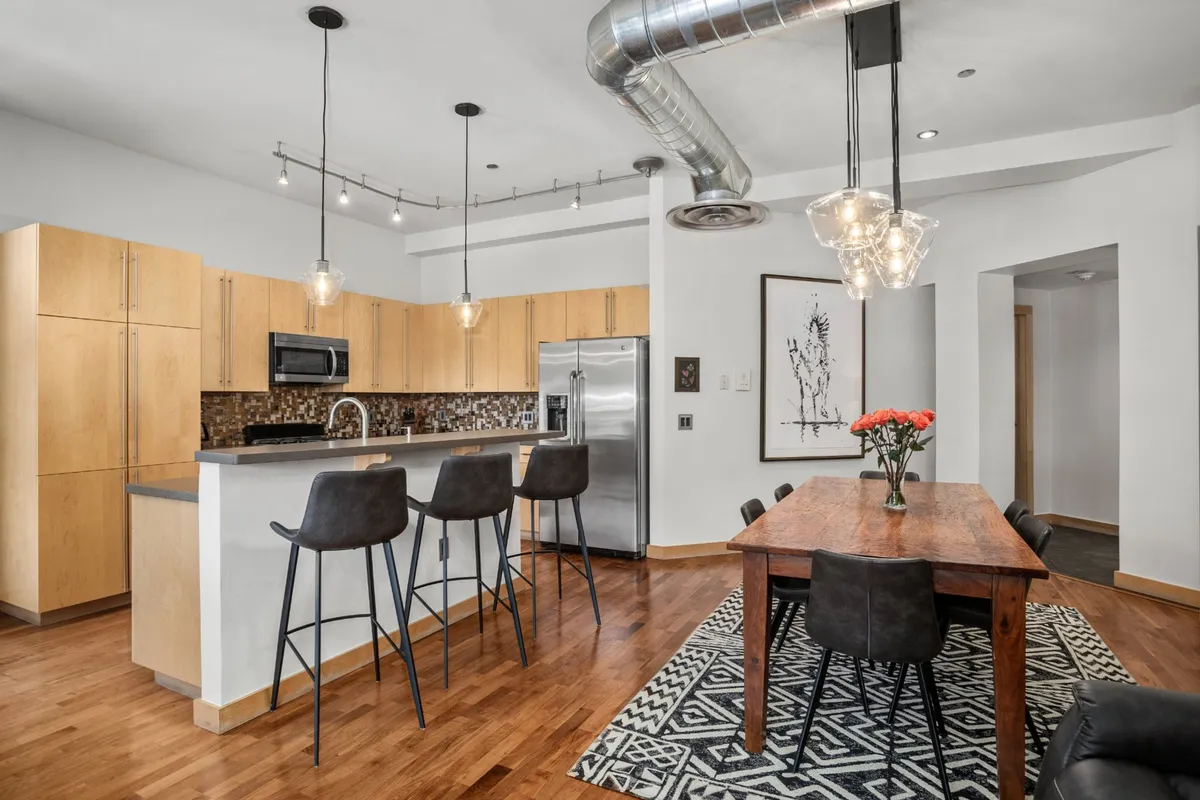 A bright, open-concept dining and kitchen area with a dining table decorated with fresh flowers, a kitchen island with bar stools, modern stainless steel appliances, and art on the wall. Large windows flood the space with natural light, and exposed metallic ductwork adds an urban touch.