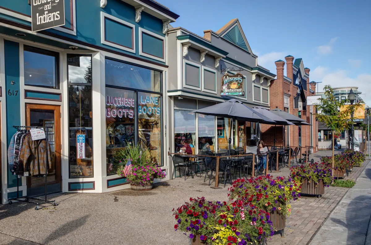 An inviting commercial streetscape featuring outdoor dining tables, colorful flower planters, and clear storefront signage. The ambiance is bright and engaging, with people enjoying food and drinks while others stroll and shop. The scene evokes a sense of community and the pleasures of outdoor dining and local discovery.