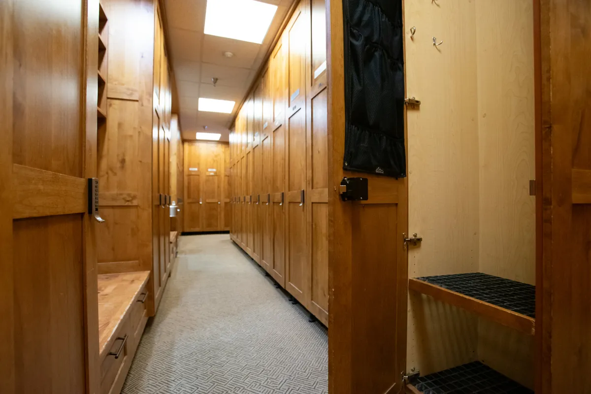 This image depicts a well-lit interior ski club space featuring wooden lockers, a carpeted floor, and an overhead skylight. The communal setting suggests a comfortable area for storing ski and snowboard gear, with space for guests to socialize and prepare for outdoor activities.