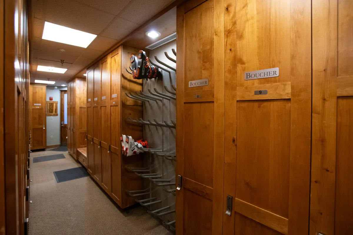 Rows of wooden lockers filled with ski equipment are shown under bright lights, with a bench for changing attire. The mood is both practical and inviting, suggesting a communal atmosphere where skiers prepare for the slopes or wind down after a day on the mountain.