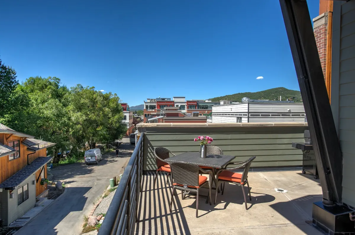 A balcony seating area overlooking charming downtown buildings, with mountains in the background. The space features comfortable chairs and a relaxed setting perfect for sipping morning coffee or enjoying evening sunsets. The view connects the luxury condo to both the lively town and nature beyond.