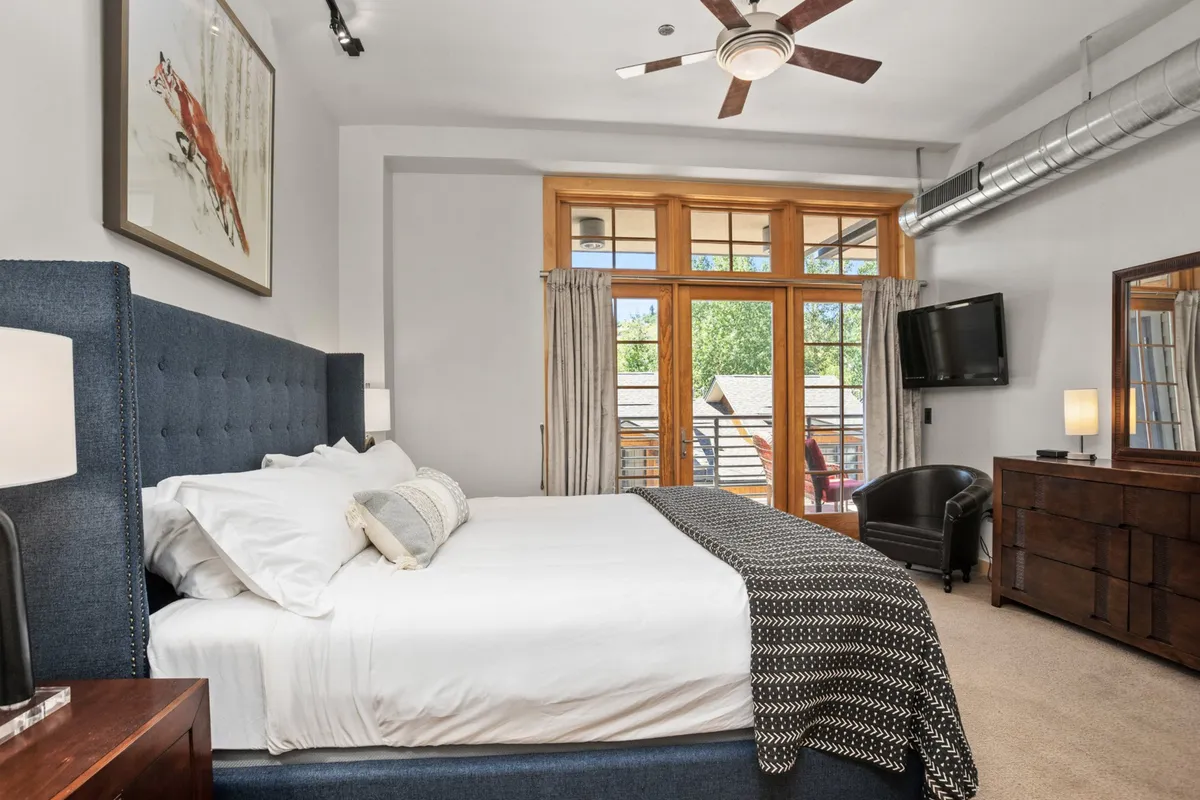 A serene, comfortable bedroom with a large bed, windows that let in natural light, a stylish dresser, an armchair, and wall artwork. The space radiates tranquility and modern comfort, ideal for restful mornings and quiet moments.