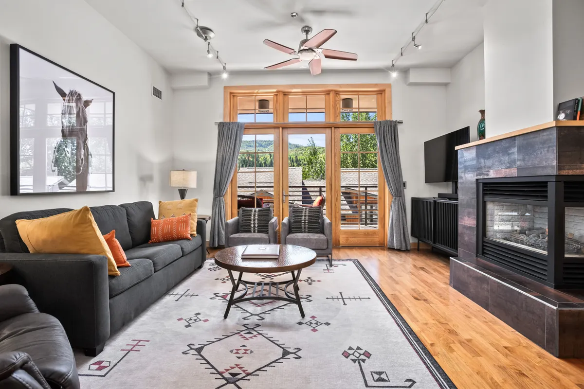 A cozy living room with large windows, a glowing fireplace, stylish artwork, and a coffee table. The inviting atmosphere suggests a warm retreat after outdoor adventures.
